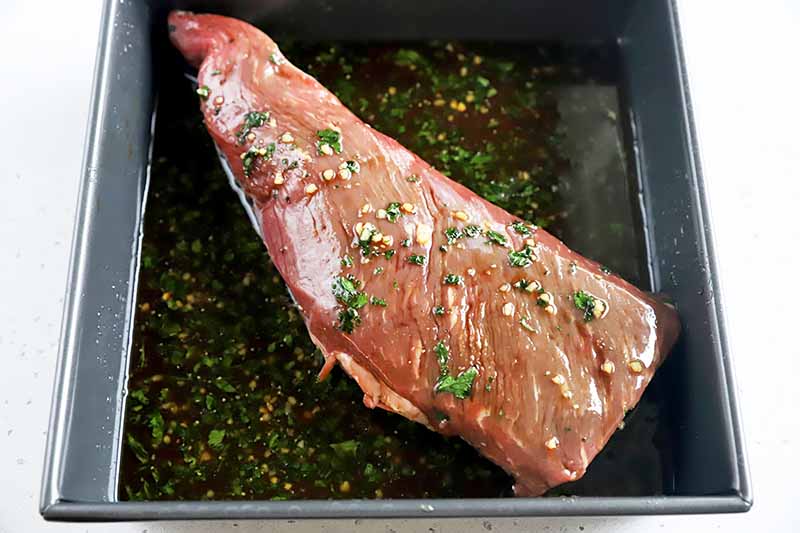 Horizontal image of a raw cut of meat marinated in a dark liquid with chopped herbs in a pan.