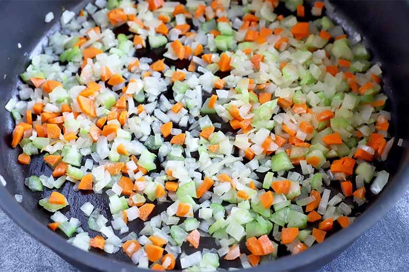 Horizontal image of chopped onion, celery and carrots cooking in oil in a pan.