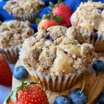 Horizontal image of baked goods in paper liners topped with crumbles on a wooden board surrounded by fresh fruit.