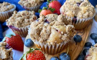 Horizontal image of baked goods in paper liners topped with crumbles on a wooden board surrounded by fresh fruit.