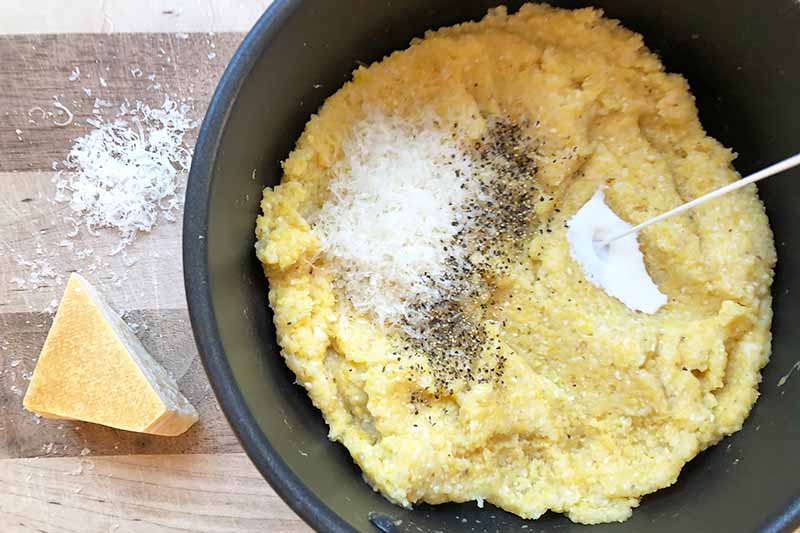 Horizontal image of cooked cornmeal in a pot with a pile of grated cheese, pepper, and a rubber spatula.