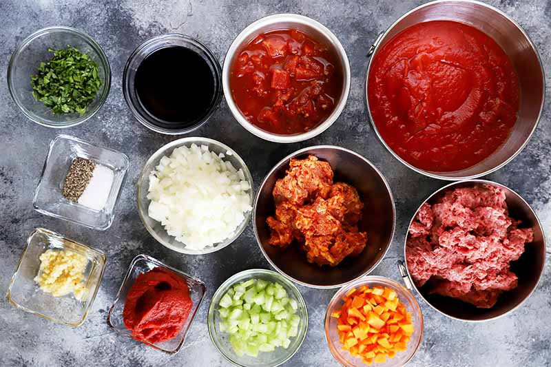 Horizontal image of many prepped ingredients in bowls to make a tomato meat sauce.
