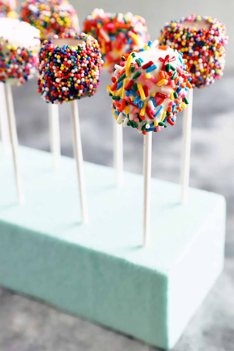 Vertical image of sprinkle and chocolate covered candies on sticks on a blue rectangular box.