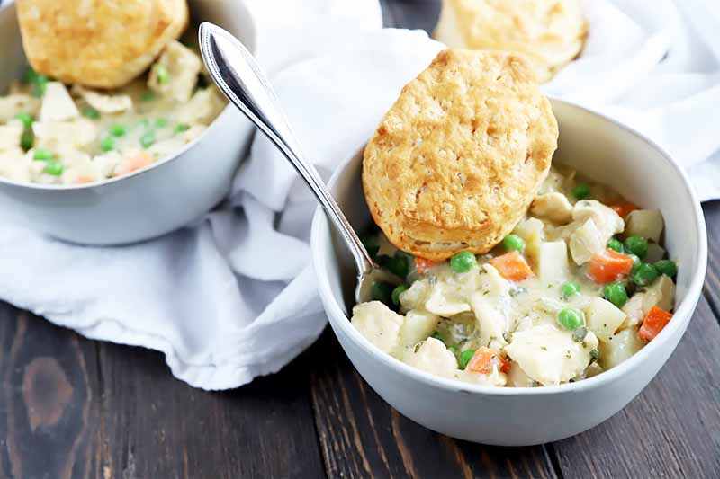 Horizontal image of two white bowls filled with a thick and creamy savory mixture of vegetables and white meat topped with biscuits.