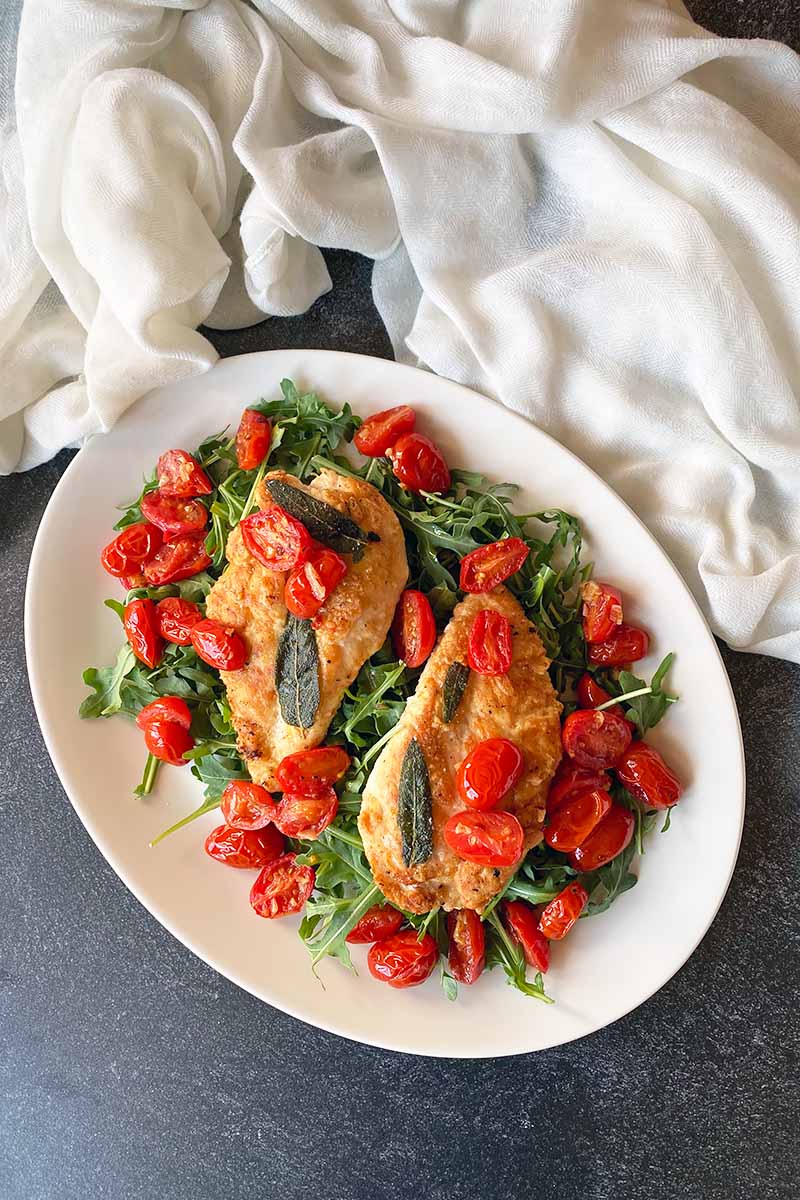 Vertical top-down image of two chicken breasts with grape tomatoes and arugula on a large white serving platter on a dark surface next to a white towel.