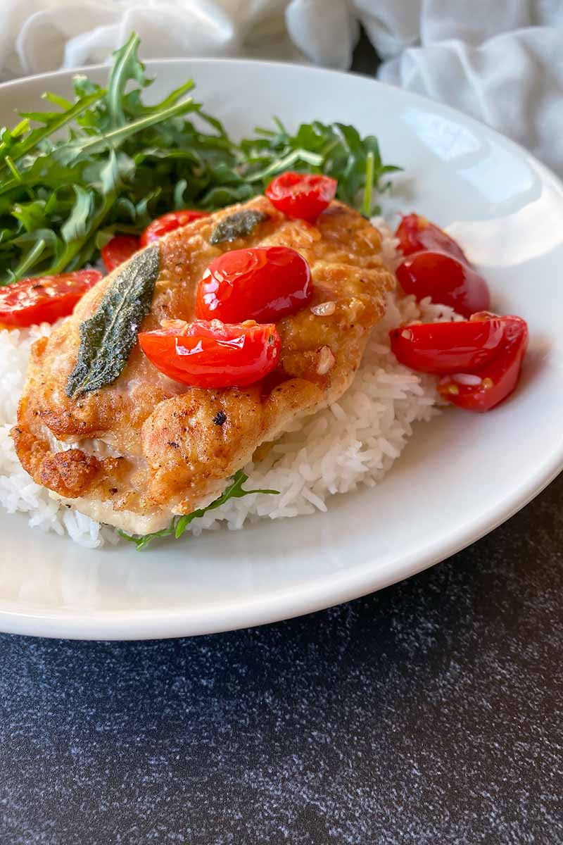 Vertical image of a cooked chicken breast with herbs and sliced tomatoes over rice on a white dish.
