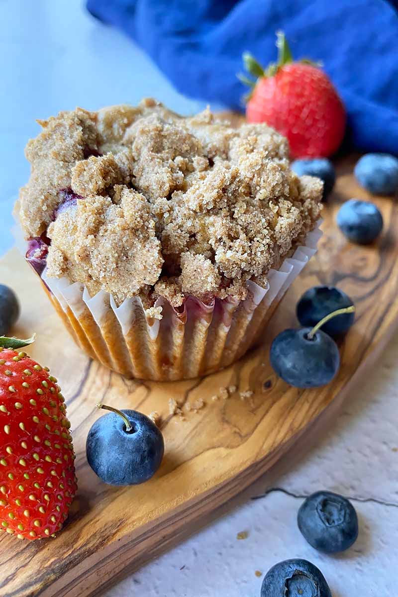 Vertical image of a streusel-topped muffin in a liner on a wooden board next to fresh fruit and a towel.