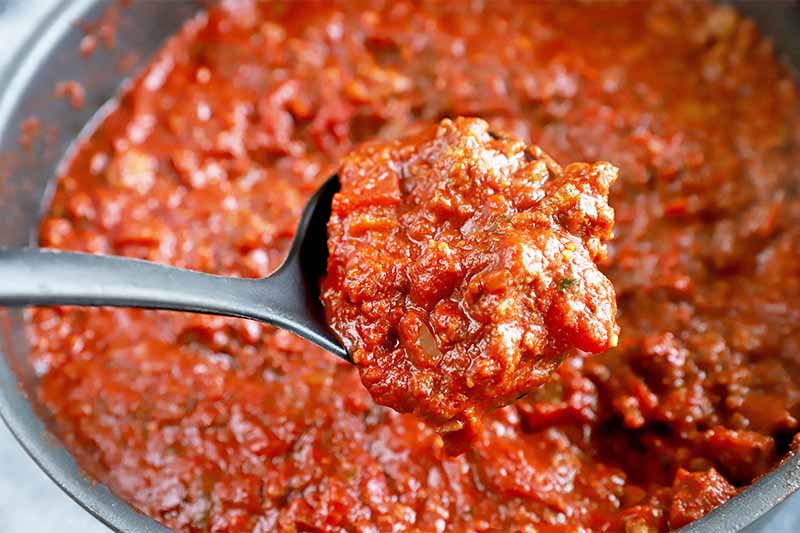 Horizontal image of a ladle holding a large amount of ragu over a pot.