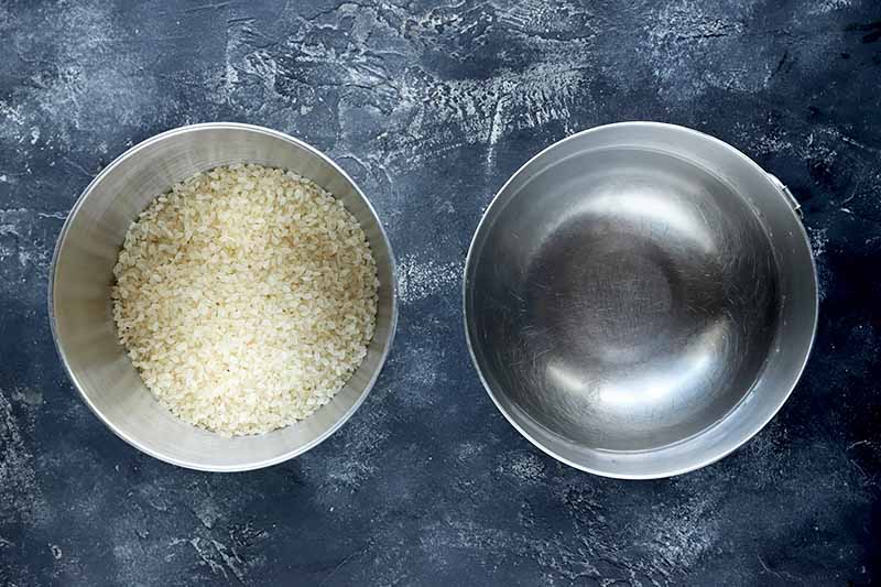 Horizontal image of a metal bowl with uncooked rice and a metal bowl with water.