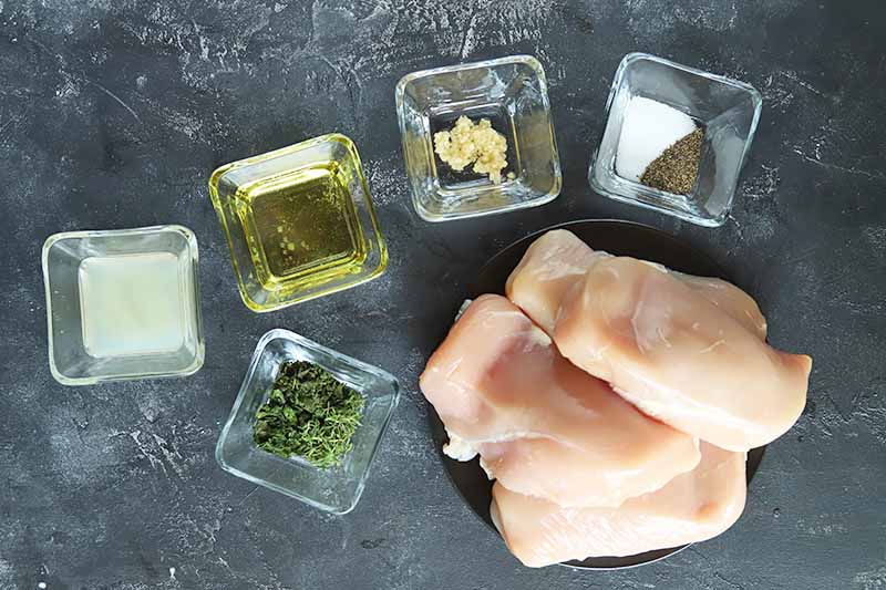 Horizontal image of raw poultry breasts and various seasonings and ingredients in square glass bowls.