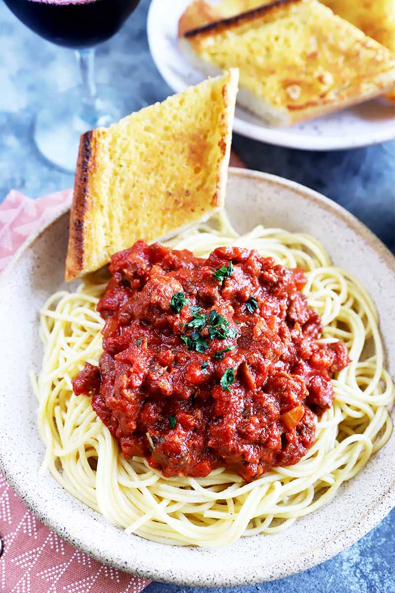 Vertical image of a plate of pasta topped with a meaty tomato ragu next to slices of garlic bread.