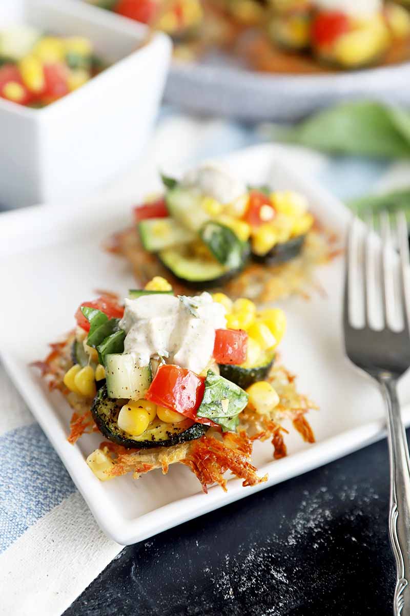 Vertical image of a white plate with two of the same fritters topped with zucchini slices, a tomato corn salad, and a dollop of yogurt next to a fork.