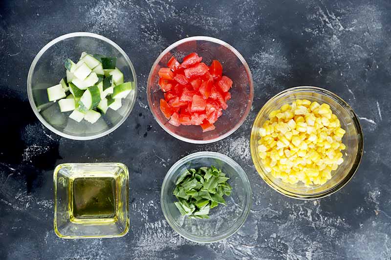 Horizontal image of assorted prepped fresh vegetables, herbs, and oil in glass bowls.