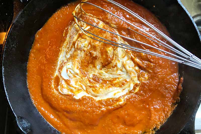 Horizontal image of whisking cream into a tomato sauce in a skillet.