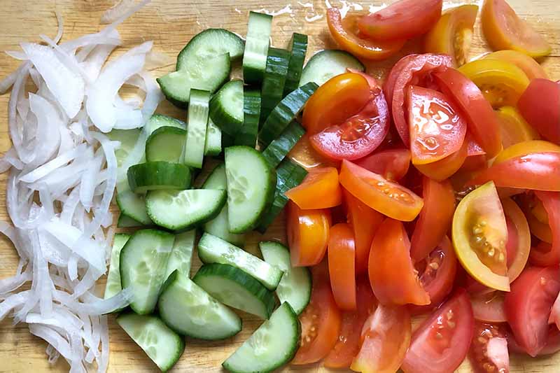 Horizontal image of slices of onions, cucumbers, and tomatoes on a wooden cutting board.