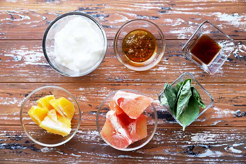 Horizontal image of cut citrus fruit, yogurt, seasonings, and basil in small glass bowls.