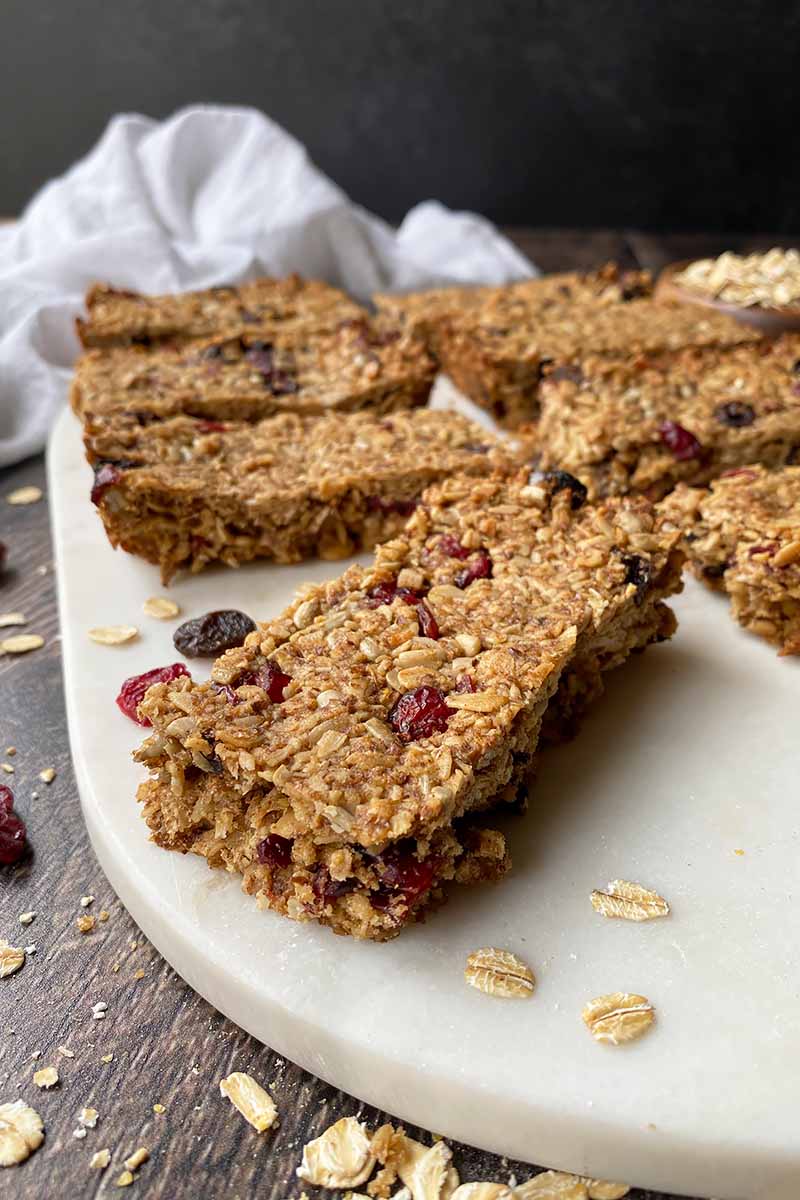 Vertical image of rows of granola bars studded with dried fruit on a white cutting board.