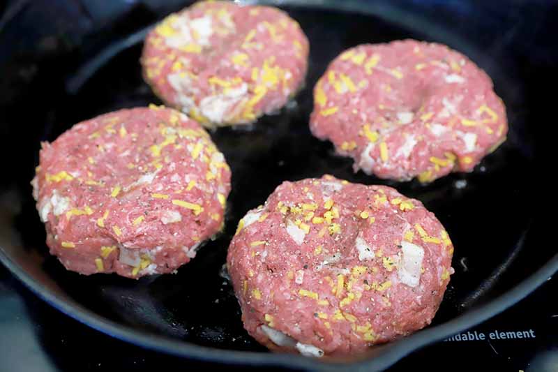 Horizontal image of cooking four raw meat patties in a cast iron skillet.