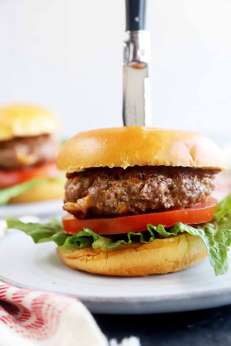 Vertical image of a knife inserted into a burger on a white plate.