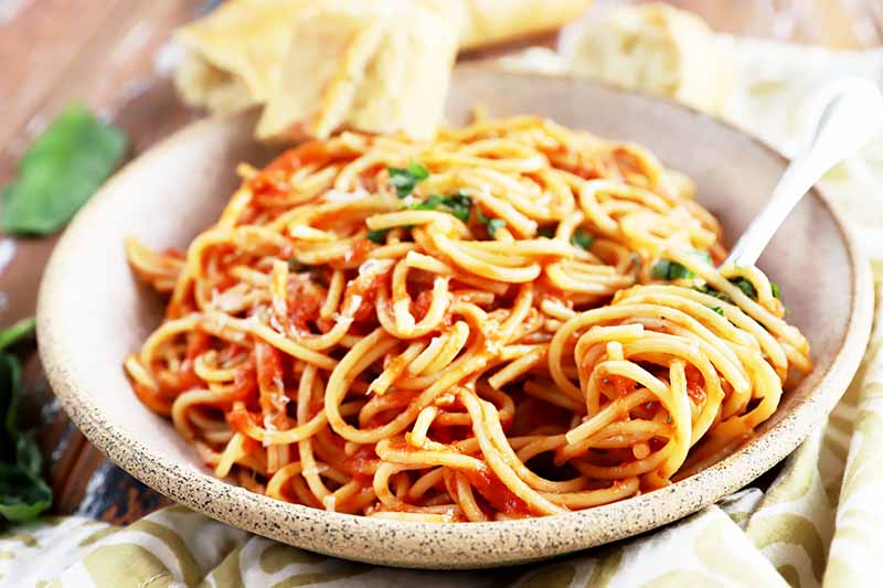 Horizontal image of a bowl full of noodles in tomato sauce with herb and cheese garnish next to a fork and towel.
