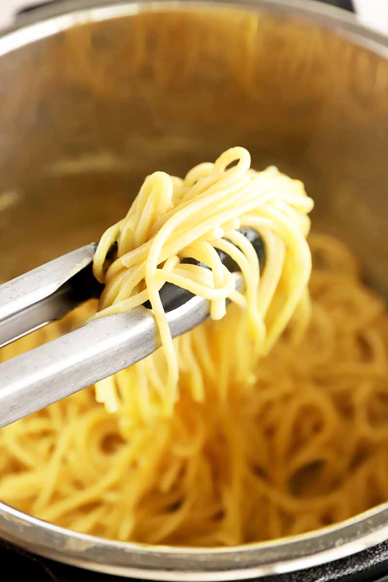 Vertical image of tweezers holding cooked pasta in a pot.