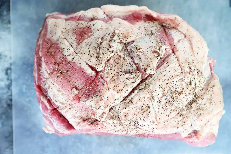 Horizontal image of a scored and seasoned large piece of raw meat on parchment paper.