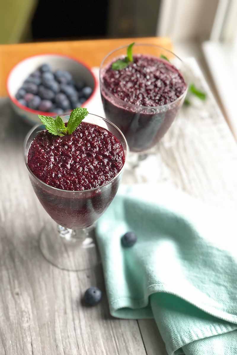 Vertical image of two glasses filled with a dark purple thick beverage next to a blue towel and a bowl of blueberries.