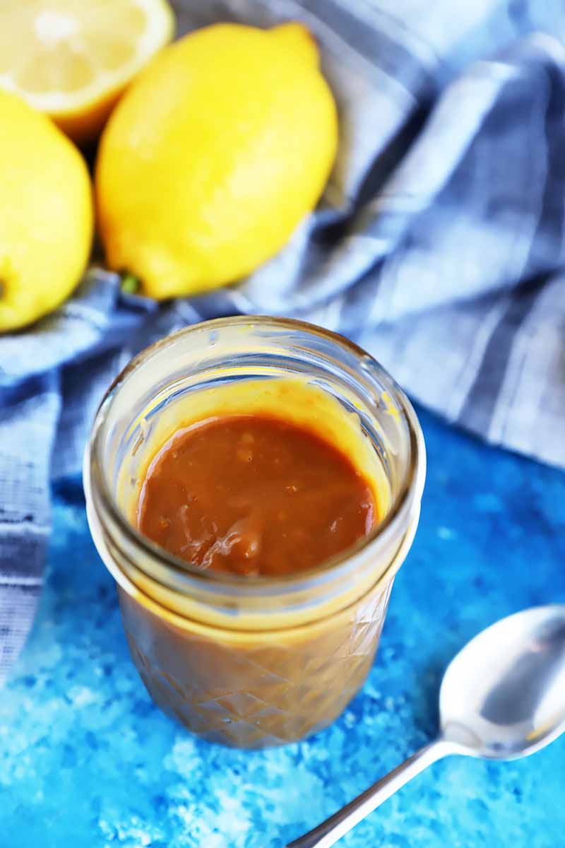 Vertical image of a mason jar filled with a dark brown thick spread next to lemons, a towel, and a spoon.