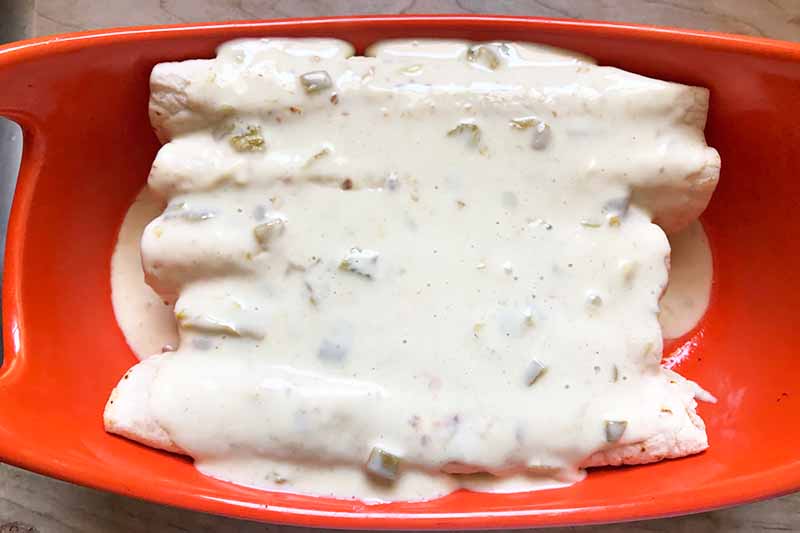 Horizontal image of unbaked enchiladas in a red dish topped with a creamy cheese sauce.