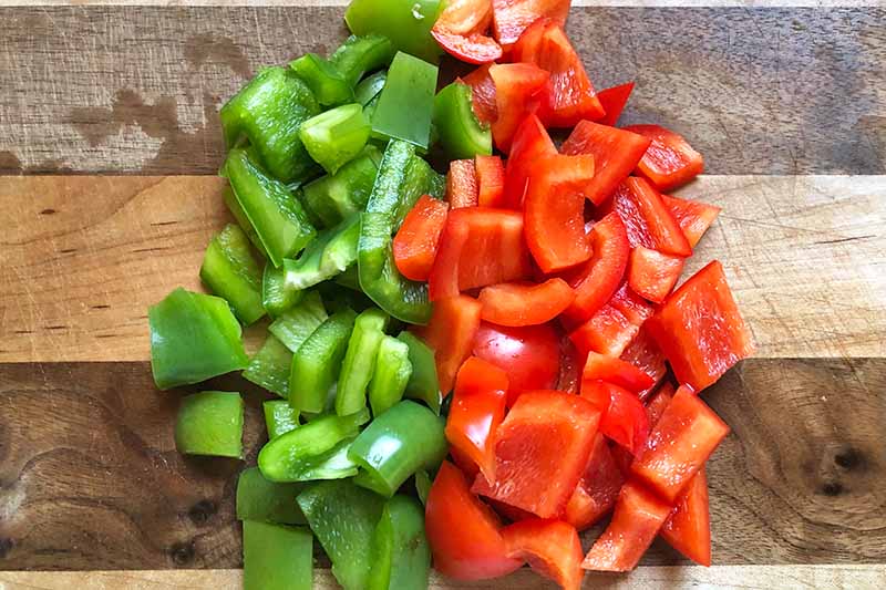 Horizontal image of green and red bell peppers chopped on a wooden cutting board.