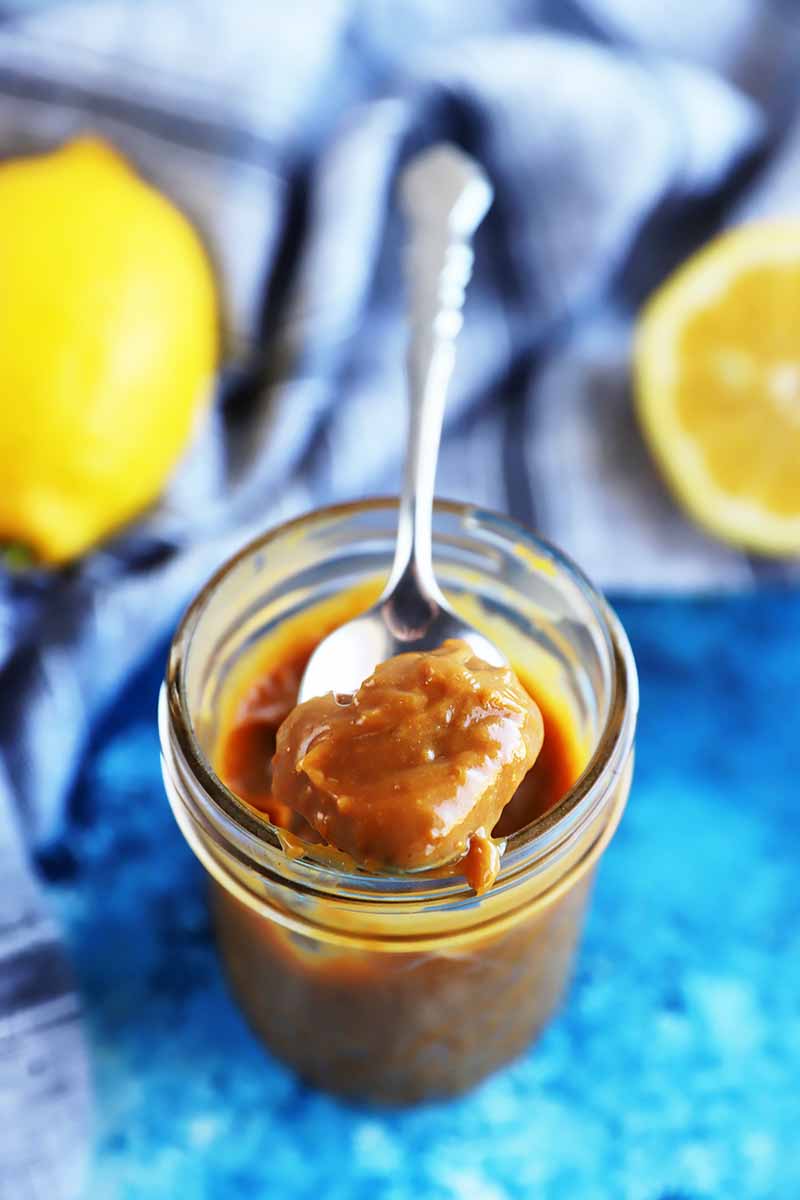 Vertical image of a spoon holding a thick spoonful of dark curd in a mason jar next to lemons.