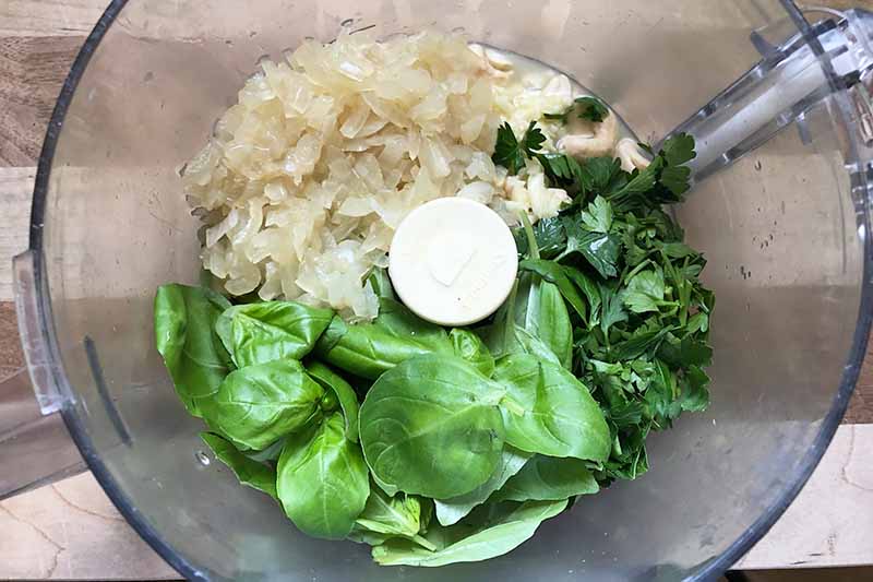 Horizontal image of cooked onions, greens, and herbs in a food processor.