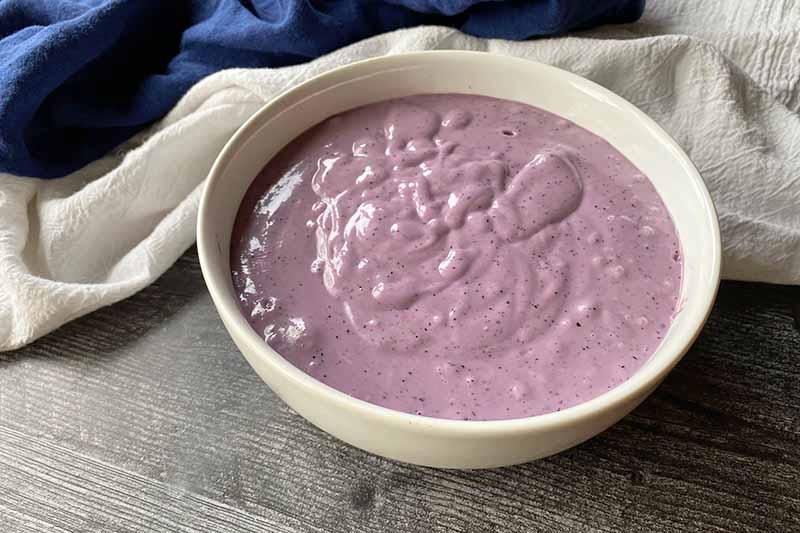 Horizontal image of a thick and smooth purple mixture in a white bowl in front of blue and white towels.