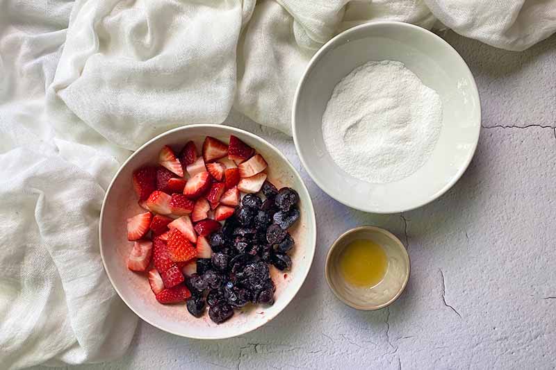 Horizontal image of a bowl of cut strawberries and cherries, a bowl of sugar, and a bowl of juice next to a white towel.