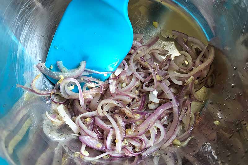 Vertical image of mixing together cooked sliced red onion and garlic in a bowl with a blue spatula.