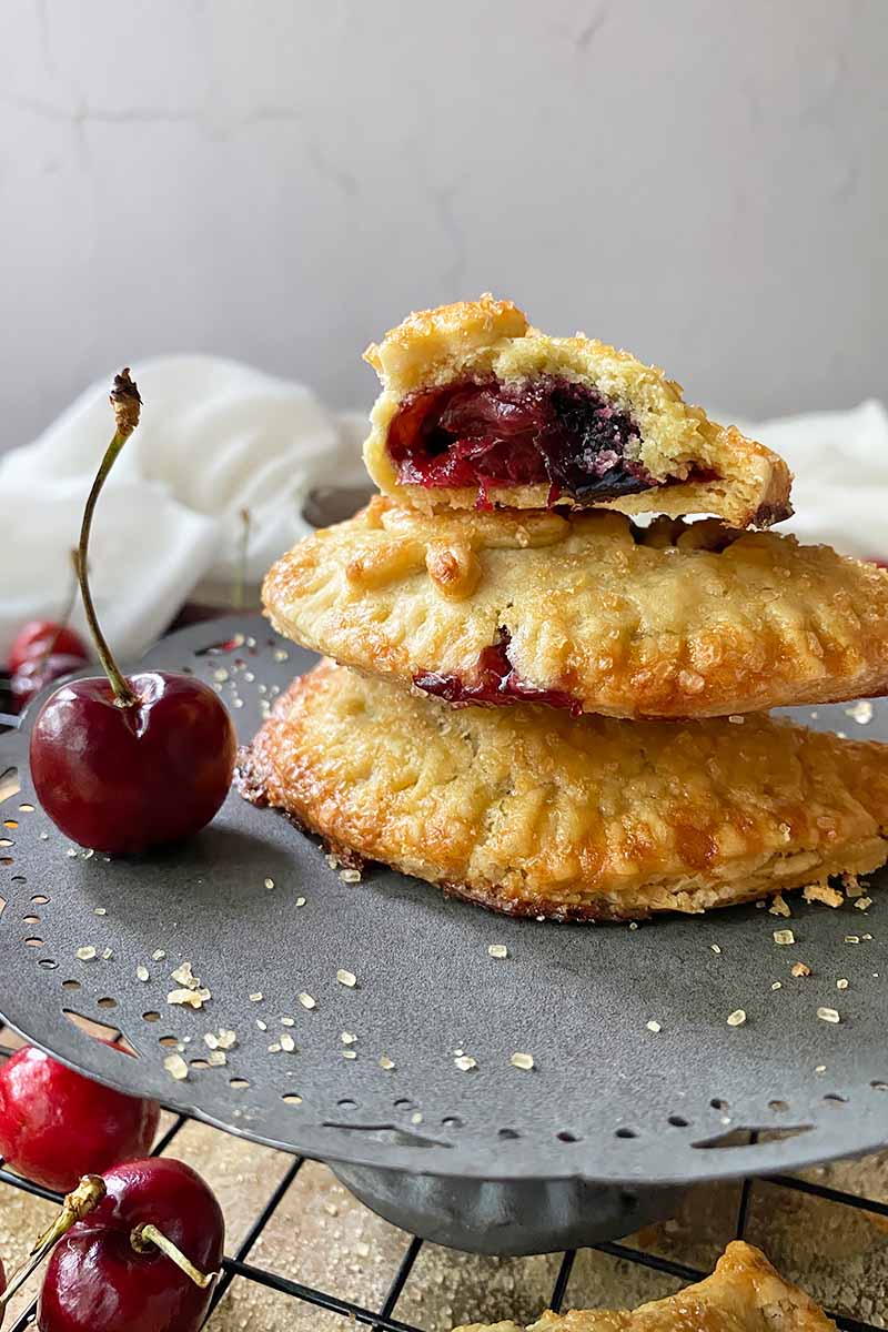 Vertical image of a stack of half-moon hand pies with a berry filling on a cake stand next to cherries.