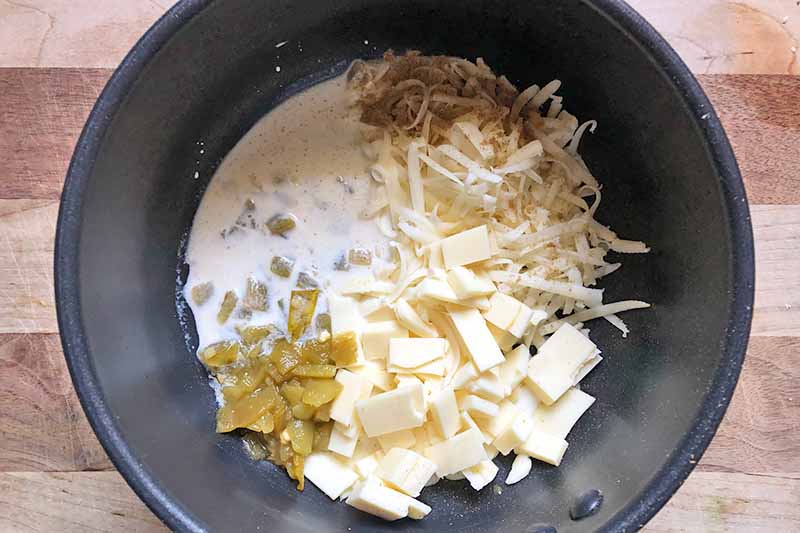 Horizontal image of assorted grated and cubed cheese, milk, and green chiles in a pot.