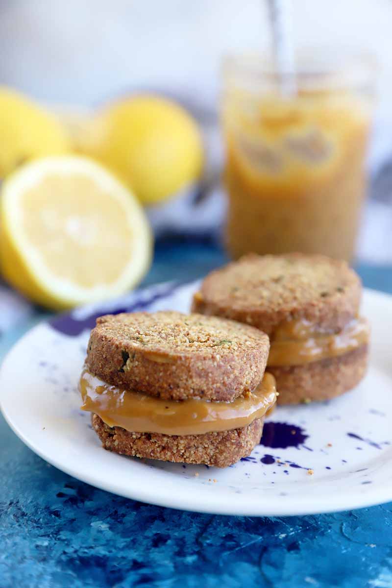 Vertical image of two cookie sandwiches filled with a light brown spread on a plate in front of citrus fruit.
