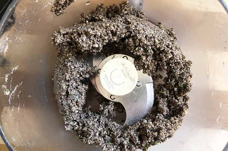 Horizontal image of a crumbly dark brown dough mixture in a food processor.