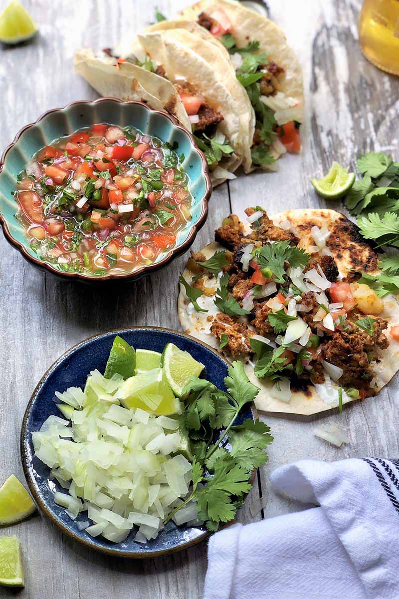 Vertical image of a bowl of pico de gallo, a bowl of chopped onions, and tacos.