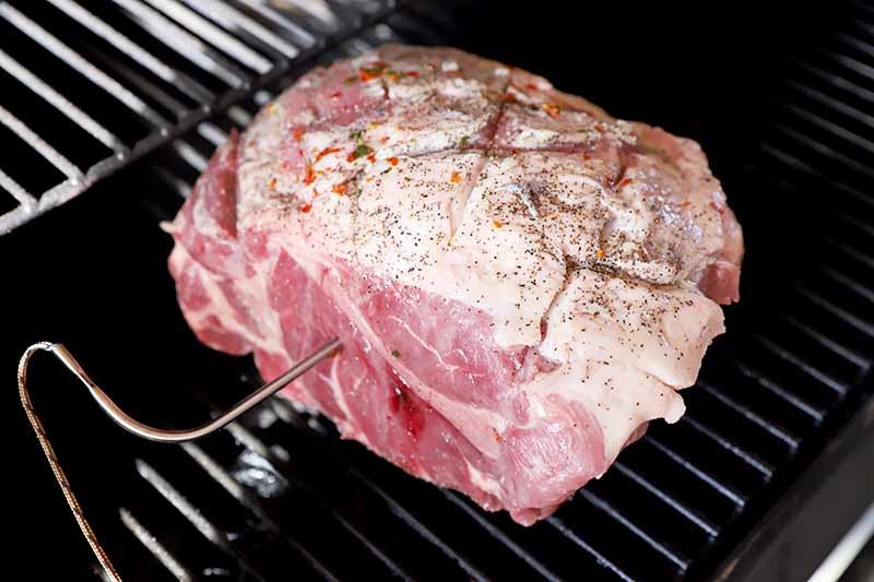 Horizontal image of a large raw chunk of scored meat on a grill held by a stake.