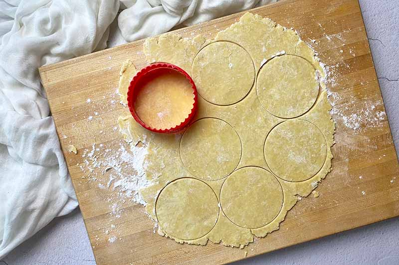 Horizontal image of cutting out circles in a flat mound of dough on a wooden cutting board.