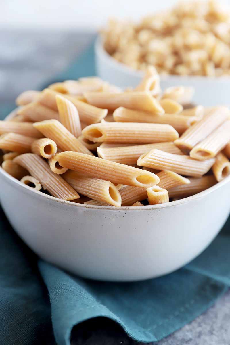 Vertical close-up image of white bowls filled with plain penne on a teal towel.
