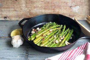 Horizontal image of green vegetables in a cast iron pan garnished with nuts next to a lemon and garlic.
