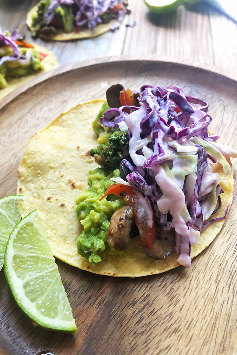 Vertical image of a flat corn tortilla topped with mashed avocado and a cabbage slaw next lime wedges on a wooden plate.