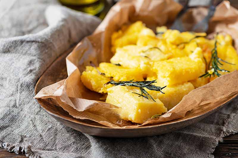 Horizontal image of fried squares of savory yellow cakes on a platter with parchment paper and fresh herbs.