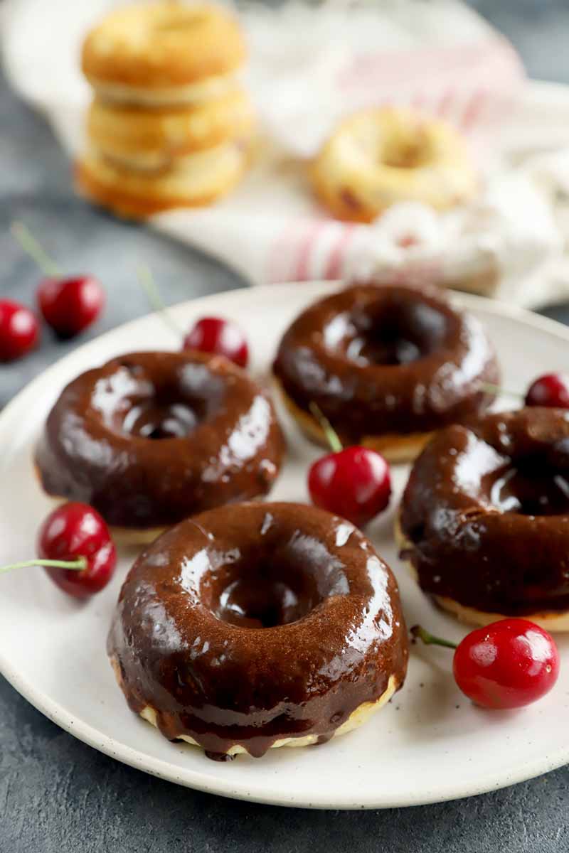 Vertical image of doughnuts covered in a chocolate glaze on a white plate surrounded by red fruit.