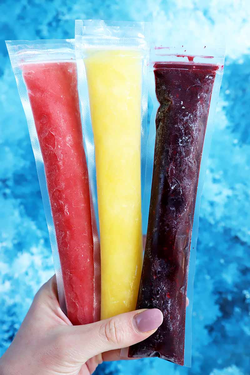 Vertical image of a hand holding three different colored ice pops over a blue surface.