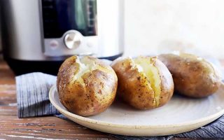 Horizontal image of three baked spuds split in the middle on a white plate next to a kitchen appliance.
