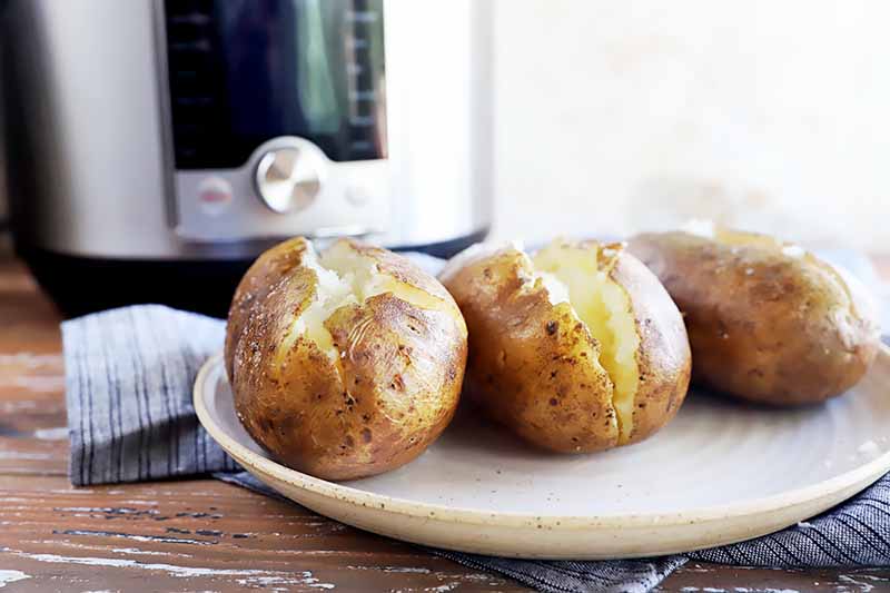 Horizontal image of three baked spuds split in the middle on a white plate next to a kitchen appliance.